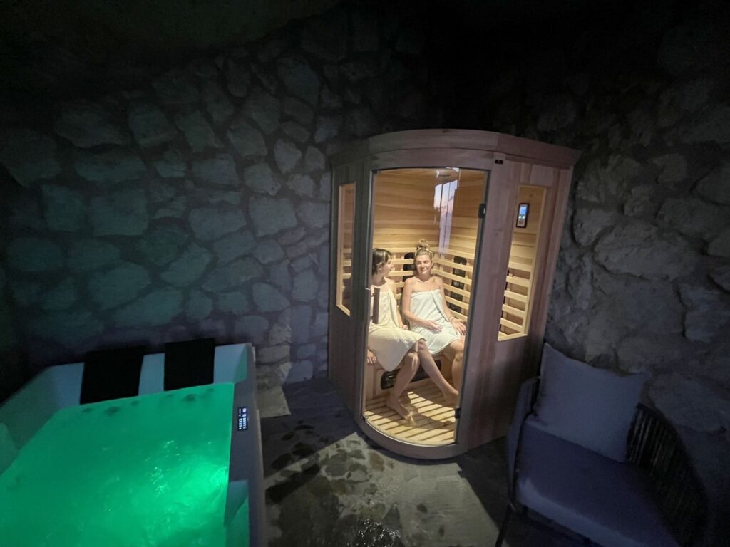 La spa nella serra: una sauna in legno con due persone sedute e una vasca idromassaggio illuminata di verde
